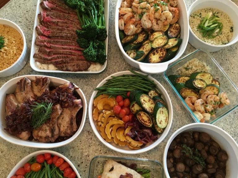 A variety of dishes on a table, including sliced beef, shrimp, sautéed vegetables, grain dishes, green beans, and a bowl of mushrooms.