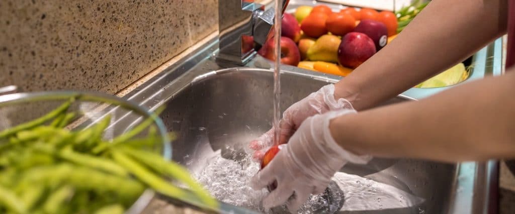 A person wearing gloves washes a red tomato under running water in a kitchen sink, ensuring food safety. Nearby, there are other fruits and vegetables on the countertop.