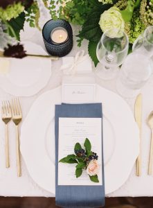 A table setting perfect for summer entertaining features a white plate with a blue napkin, menu, and floral arrangement on top. Surrounding it are gold utensils, a candle in a blue holder, wine glasses, and greenery.