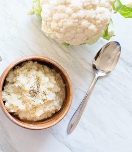 A wooden bowl of cauliflower rice garnished with black pepper, a stainless steel spoon, and a whole cauliflower on a white marble surface—an essential for healthy eating and shedding those pandemic pounds.