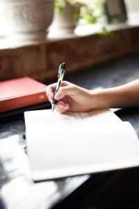 A person writes in a notebook with a pen, while a red book and potted plants are visible in the background, possibly jotting down successful strategies for healthy eating.