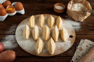 A wooden board with uncooked dumplings, surrounded by ingredients including eggs, flour, and potatoes on a rustic wooden table, showcases the essence of Hispanic traditional foods.