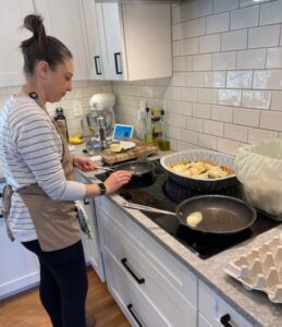 A woman wearing a striped shirt and apron cooks on a stovetop in a modern kitchen with white cabinetry and tile backsplash, reminiscent of the stylish setups often seen in Seattle nonprofits. Various kitchen items, including pans and food, are spread on the counter.