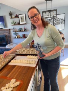 A woman stands in a kitchen, smiling at the camera while preparing pastries on a countertop. She holds a bag of flour in her hand, with trays of rolled dough in front of her. Her passion for baking extends beyond her home, as she frequently volunteers with Seattle nonprofits to share her culinary skills.