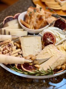 A platter with assorted cheeses, nuts, and sliced blood oranges—a nod to 2023 food trends—accompanied by bread pieces in a separate bowl.