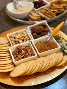 A platter with an assortment of crackers, almonds, pecans, dried fruit, and a dish of marmalade, accompanied by a cheese knife—a perfect representation of 2023 food trends. Another plate of assorted snacks is visible in the background.