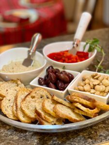 A platter with sliced bread, olives, marcona almonds, roasted red peppers, and a bowl of hummus is arranged on a countertop, reflecting 2023 food trends.