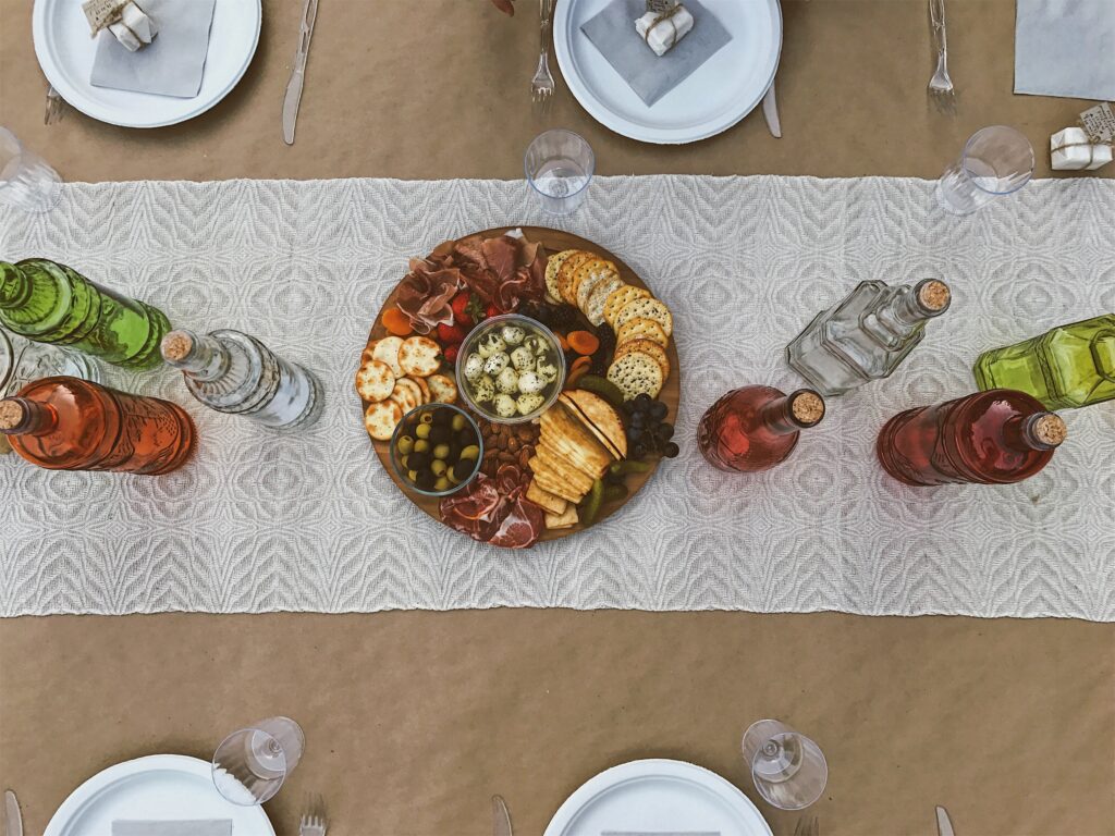 A charcuterie board with assorted meats, cheeses, crackers, and olives sits at the center of a table adorned with a lace runner, surrounded by glass bottles and plates set for a meal—a perfect display of 2023 food trends.