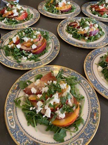 Plates of salad with arugula, sliced peaches, and crumbled cheese are arranged on a table. Fun fact about Chef Stacey O'Farrell: she's not only known for her gourmet salads but also for an unforgettable cherry pie.