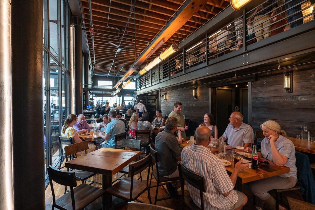 A group of people dining inside a modern, rustic-style restaurant with wooden decor and an open ceiling. Tables are arranged closely, and some diners are engaged in conversation while others eat, enjoying one of the finest Bellingham restaurants that perfectly blends cozy charm with contemporary appeal.