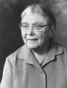 A black and white portrait of an elderly woman with glasses, wearing a light-colored jacket, and hair pulled back. She is smiling slightly and looking to her left.