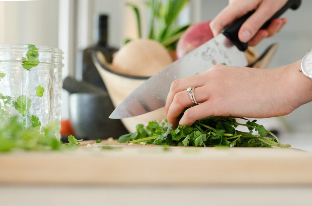 A person's hands are seen chopping fresh green herbs on a cutting board with a knife. A jar and a bowl of vegetables are in the background.