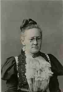 Black and white portrait of an older woman wearing glasses and a high-collared dress with lace detailing and puffy sleeves, her hair is styled up with a bow.