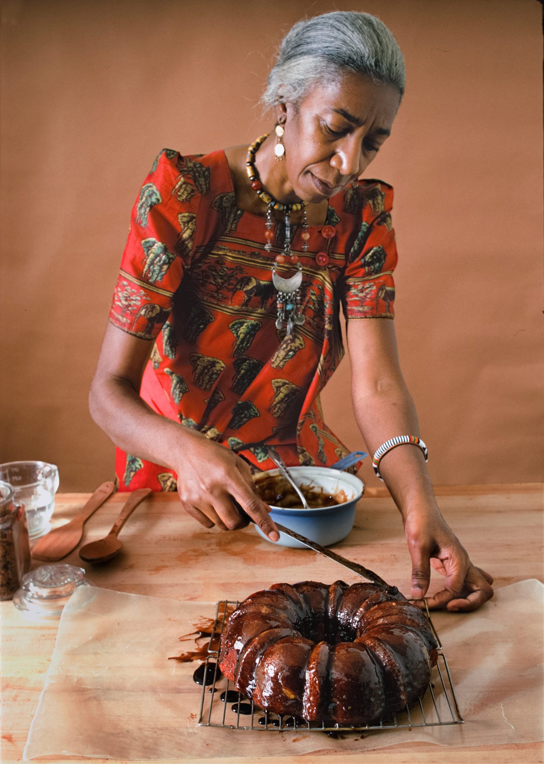 An elderly woman in a red dress decorates a Bundt cake with icing on a cooling rack, using a spoon from a nearby bowl. Kitchen utensils and ingredients are on the table.