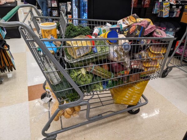 A grocery cart filled to the brim with various food items—including vegetables, canned goods, bread, meat, and packaged products—sits in a store aisle on a tiled floor. Perfect for home entertaining and mastering hospitality etiquette.