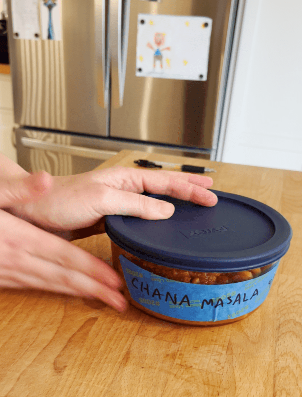 Hands placing a container labeled "Chana Masala" on a wooden countertop, showcasing simple hospitality etiquette. The container has a blue lid, and a refrigerator with a child's drawing is visible in the background, highlighting the charm of in-home entertaining.