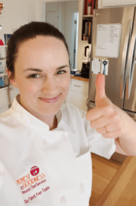 A person in a white chef's uniform is smiling and giving a thumbs up in a kitchen, with a refrigerator and a whiteboard visible in the background, embodying simple hospitality etiquette.