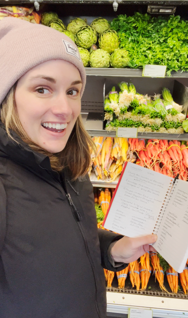 A person wearing a beanie and jacket smiles while holding a notebook in front of a grocery store produce section with various vegetables, perhaps noting ingredients for some simple hospitality etiquette tips.