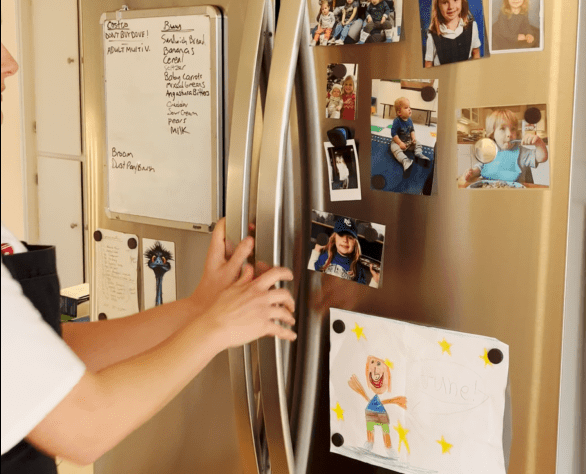 Person opening a stainless steel refrigerator door covered with family photos, children's drawings, and a whiteboard with handwritten notes—a perfect blend of personal touches that set the stage for warm in-home entertaining.