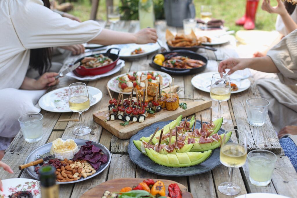 A group of people seated around a wooden picnic table covered with various dishes, such as fruit, skewers, cheese, and wine glasses with white wine, enjoying a meal together outdoors. The scene captures the essence of simple hospitality as they savor the joys of in-home entertaining.