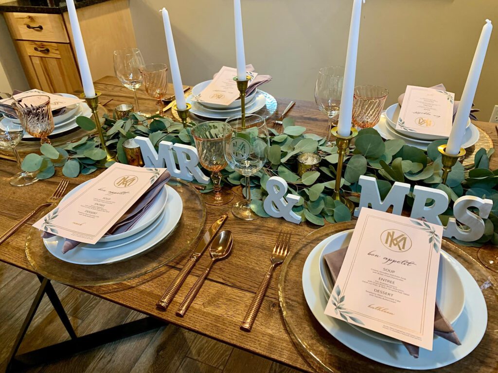 Elegant table setting for a wedding, featuring "MR & MRS" letters, several place settings with menus, glasses, and greenery decor. Tall white candles are placed in the center of the table.