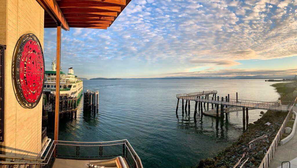 A docked ferryboat is next to a building with a large decorative red sign. An empty pier extends into the calm water under a partly cloudy sky, offering an idyllic setting perfect for anniversary date ideas.