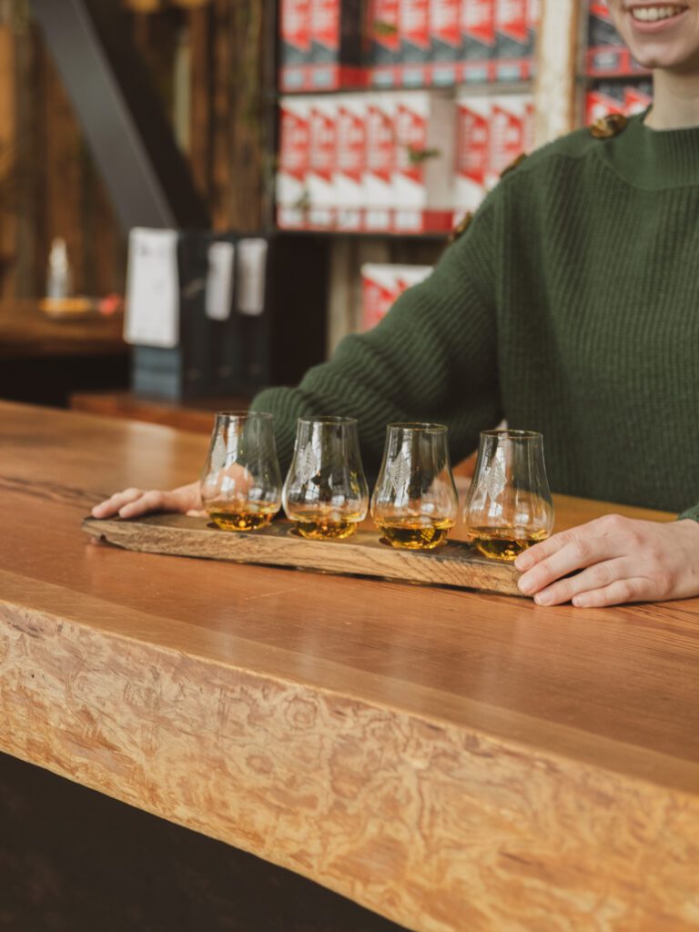 A person in a green sweater holding a wooden tray with four glasses of Westland Distillery whiskey on it at a bar counter.