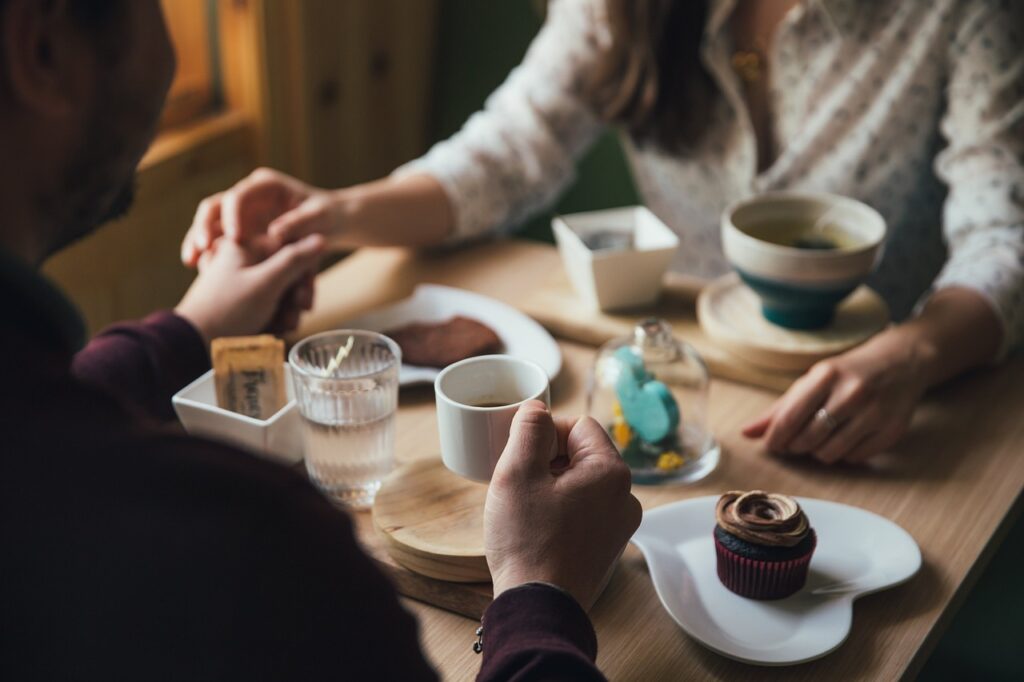 Two people sit at a table holding hands, with assorted desserts and beverages in front of them, reminiscing about their fun cooking class earlier that day.