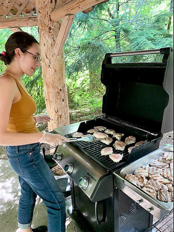 A person wearing a yellow tank top and jeans grills multiple pieces of meat, including sizzling burgers, on a gas barbecue under a wooden canopy in a forested area.