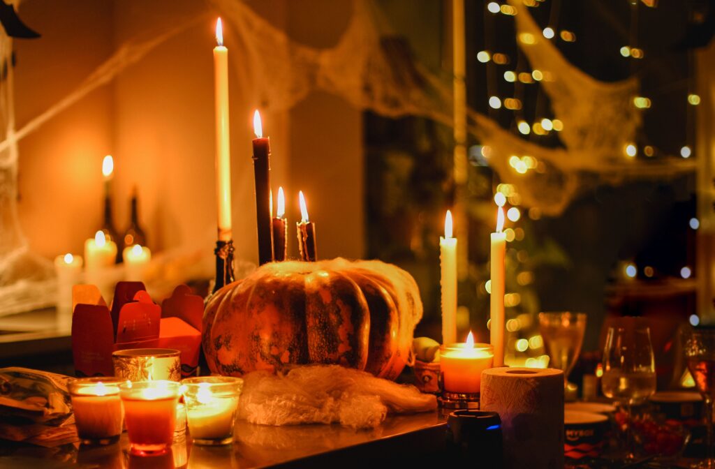 A dimly lit table decorated with lit candles, a large pumpkin, cobwebs, and string lights creates a warm, festive atmosphere perfect for Halloween.