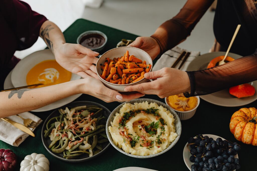 Two friends at a Friendsgiving dinner pass a bowl of roasted carrots across a table set with various dishes, including mashed potatoes, green beans, grapes, and soup.