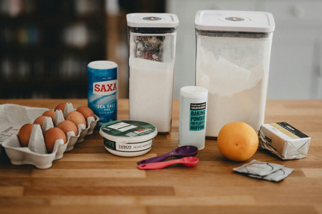 A variety of baking ingredients, including eggs, salt, sugar, flour, baking powder, mascarpone cheese, an orange, butter, and measuring spoons with labels to decode are arranged on a wooden countertop.