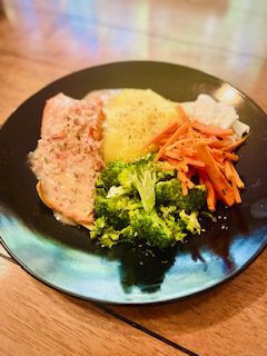 A black plate with baked salmon, mashed potatoes, steamed broccoli, and julienned carrots, artfully presented by Chef Jane Hall on a wooden table.