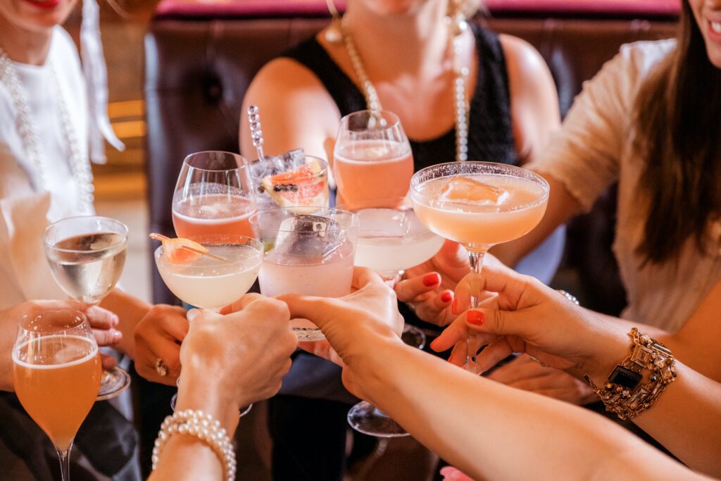 A group of people are toasting with various cocktails and drinks at a social gathering, some savoring their mocktails as they embrace Dry January. The background shows a dimly lit, cozy setting.