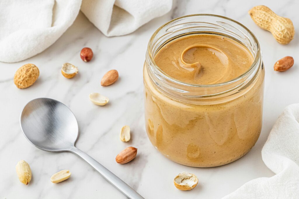 An open jar of peanut butter on a marble surface, surrounded by peanuts in shells and a metal spoon.