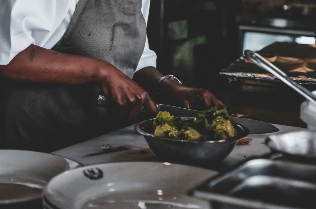 A person wearing an apron is chopping vegetables on a cutting board, with a bowl of leafy greens and plates on a kitchen counter, embodying the spirit of black excellence inspired by chefs like Edna Lewis.