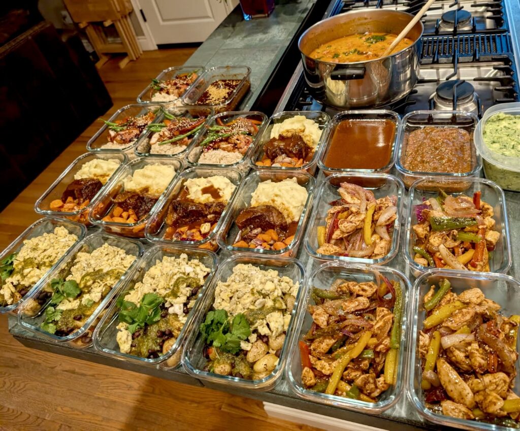 Glass meal prep containers filled with various cooked dishes, including vegetables, proteins, and sauces, arranged on a kitchen counter highlight the convenience of Personal Chef Services next to a pot on a stove.