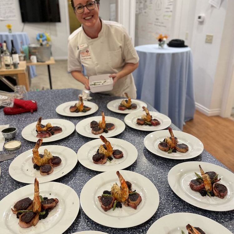 A chef in a white coat stands behind a counter displaying multiple plates of a gourmet shrimp and vegetable dish. The setting appears to be a catered event or cooking class, showcasing exceptional hospitality etiquette.