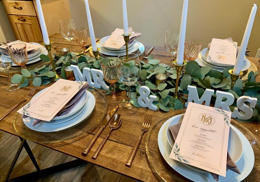 Elegant table setting for a wedding, featuring "MR & MRS" letters, several place settings with menus, glasses, and greenery decor. Tall white candles are placed in the center of the table.