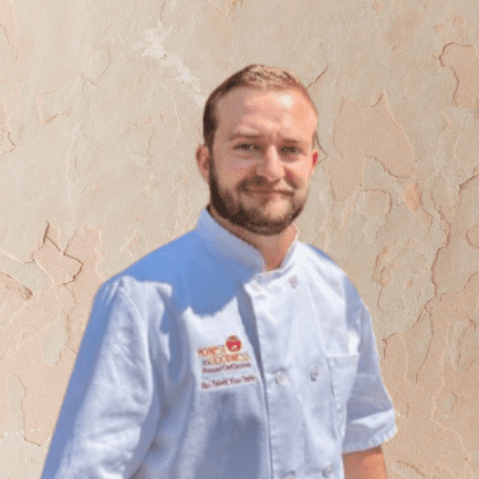 A man with a beard wearing a white chef’s coat stands in front of a beige textured wall, ready to showcase his Personal Chef Services.