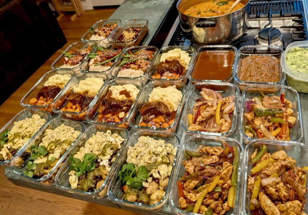 Glass meal prep containers filled with various cooked dishes, including vegetables, proteins, and sauces, arranged on a kitchen counter highlight the convenience of Personal Chef Services next to a pot on a stove.