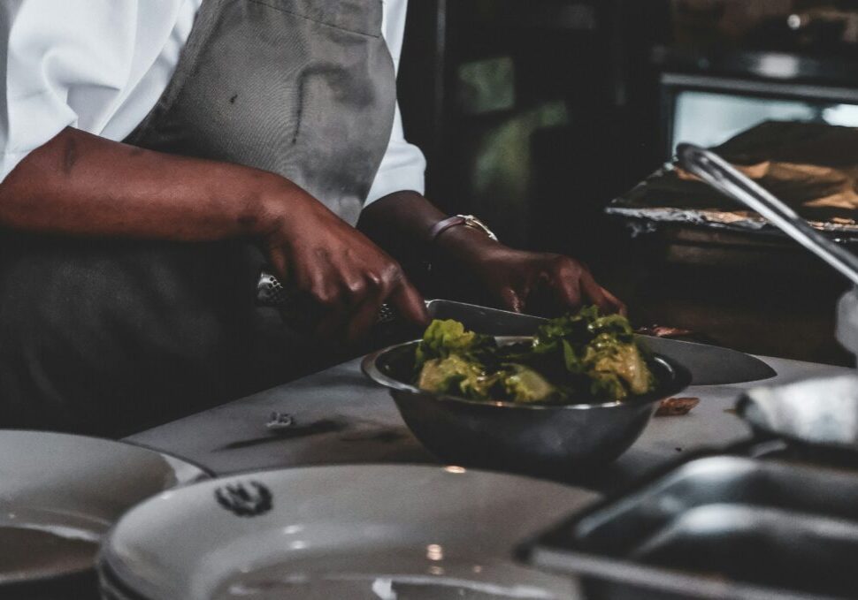 A person wearing an apron is chopping vegetables on a cutting board, with a bowl of leafy greens and plates on a kitchen counter, embodying the spirit of black excellence inspired by chefs like Edna Lewis.