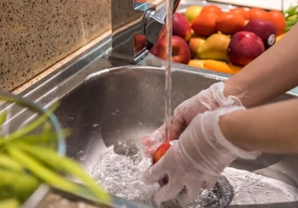 A person wearing gloves washes a red tomato under running water in a kitchen sink, ensuring food safety. Nearby, there are other fruits and vegetables on the countertop.