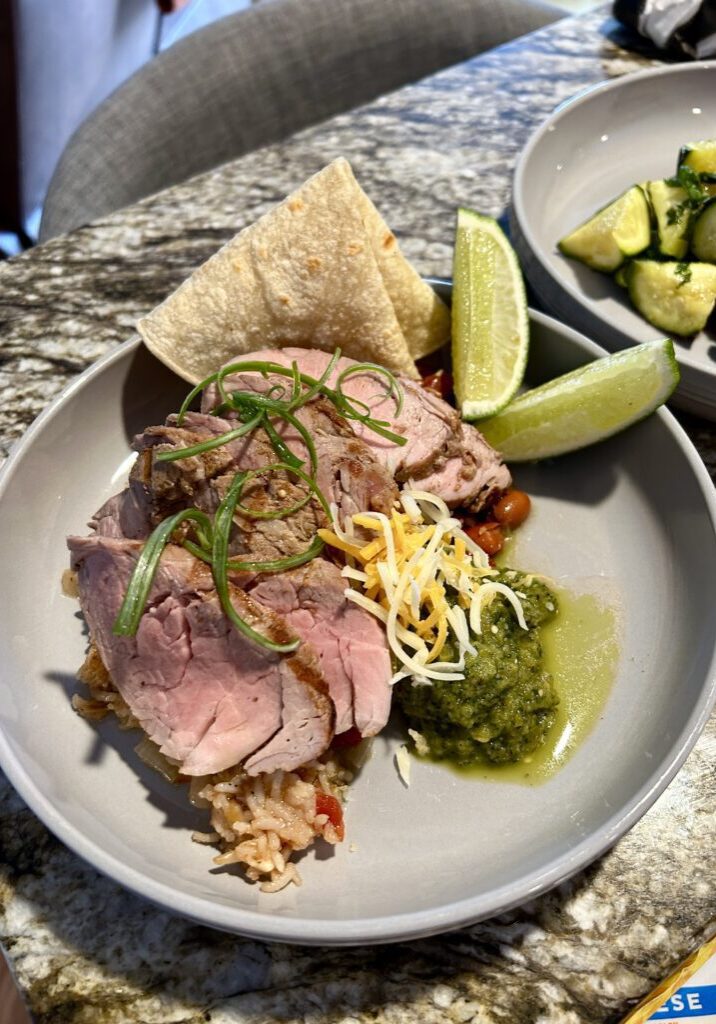 A plate with sliced roast pork on rice, shredded cheese, green salsa, pinto beans, tortilla, lime wedges, and a side of cucumber salad—perfectly crafted by our Personal Chef Services for a memorable dining experience.