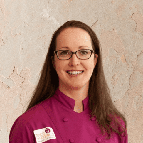 A woman with long brown hair wears glasses and a magenta chef coat, standing in front of a textured beige wall, ready to offer her Personal Chef Services.