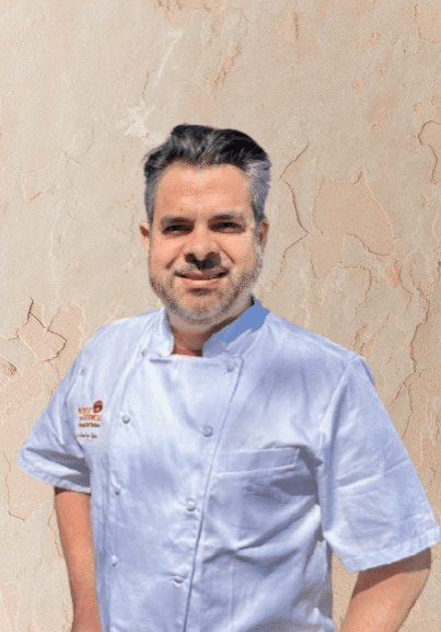 A man with dark hair and a beard, wearing a light blue chef's coat, stands in front of a textured beige wall, representing professional Personal Chef Services.