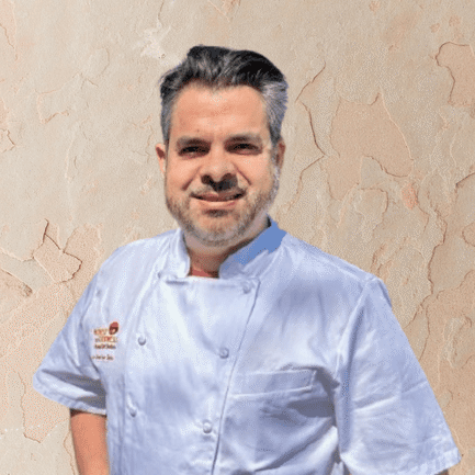 A man with dark hair and a beard, wearing a light blue chef's coat, stands in front of a textured beige wall, representing professional Personal Chef Services.