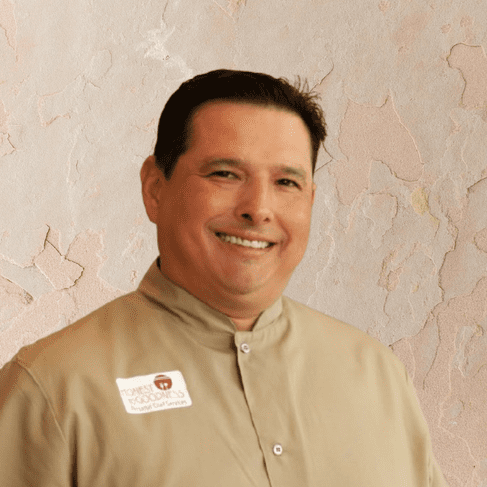 Smiling man wearing a beige button-up shirt and a name tag stands in front of a textured, light-colored wall, ready to offer exceptional Personal Chef Services.