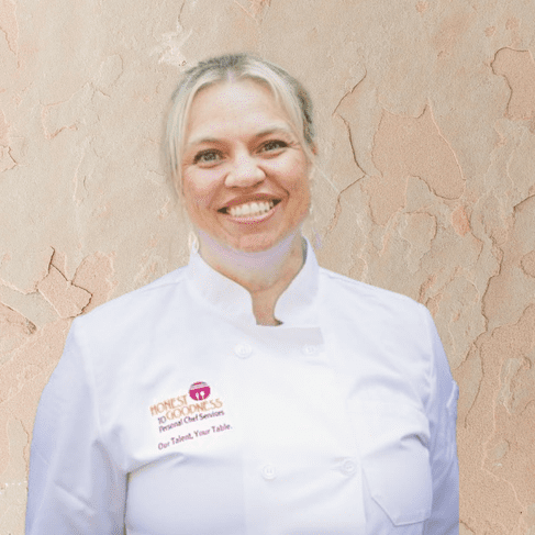 A woman wearing a white chef jacket stands smiling in front of a textured beige wall, ready to offer her Personal Chef Services.