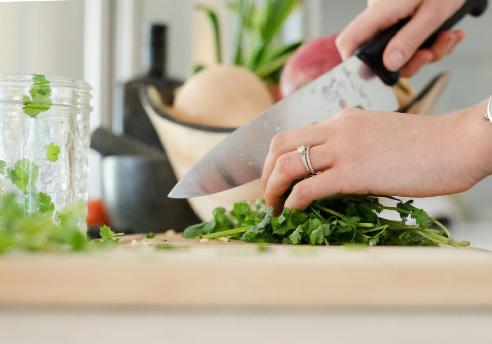 A person's hands are seen chopping fresh green herbs on a cutting board with a knife. A jar and a bowl of vegetables are in the background.